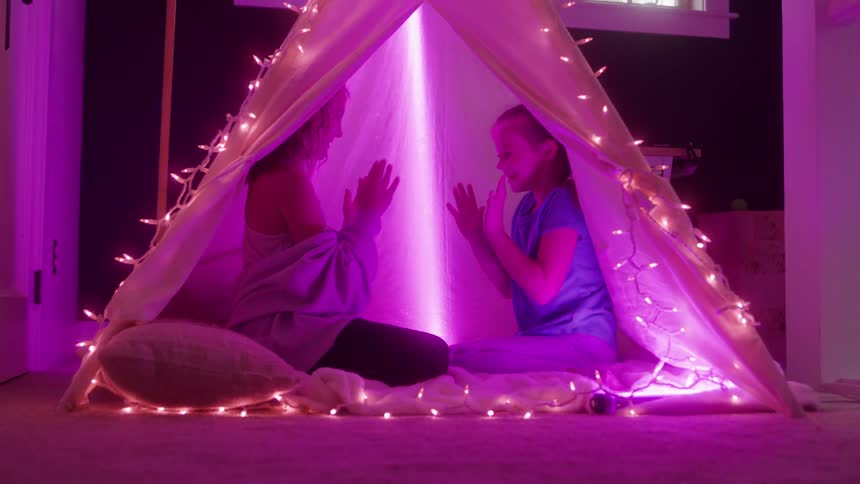 Two kids sit together playing a hand clapping game inside a string lit blanket fort