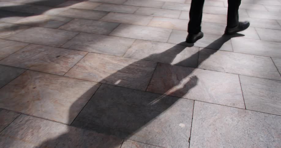 A lowangle view of a persons legs dressed in black pants and shoes walking across a patterned stone pavement while reaching a car parked
