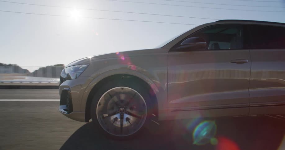 A modern gold colored suv drives quickly down a sunny coastal road with a bright lens flare and a woman standing in the distance