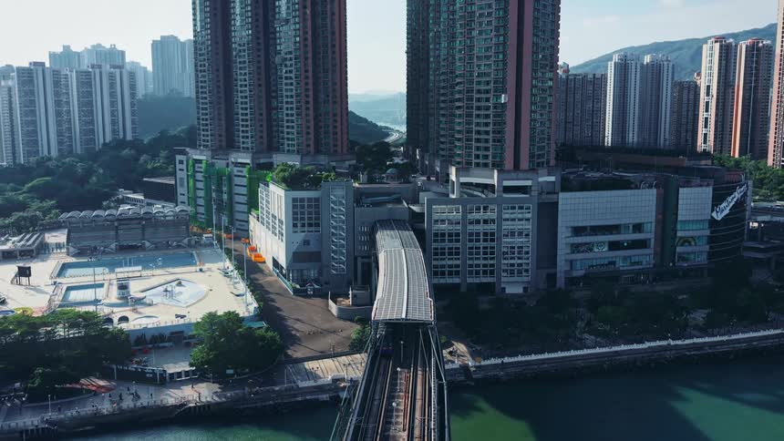 An aerial shot begins directly above a train traveling across a bridge over a body of water then slowly rises to reveal the track leading into two massive closely positioned residential towers surrounded by a dense urban environment
