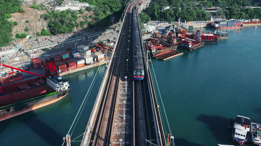 An aerial view shows a train crossing a long bridge over a harbor with cargo ships and docks connecting a residential area to a hillside and city