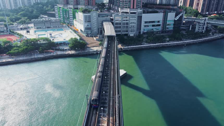 An aerial view follows a commuter train as it crosses a bridge over calm turquoise water smoothly approaching a station nestled among tall city buildings