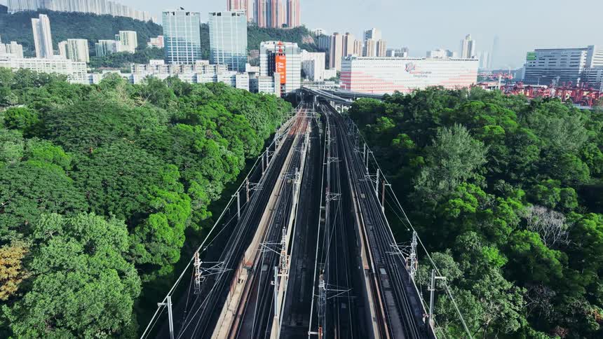 An aerial view shows multiple parallel railway tracks cutting through a dense green forest leading toward a modern city skyline and industrial port in the background