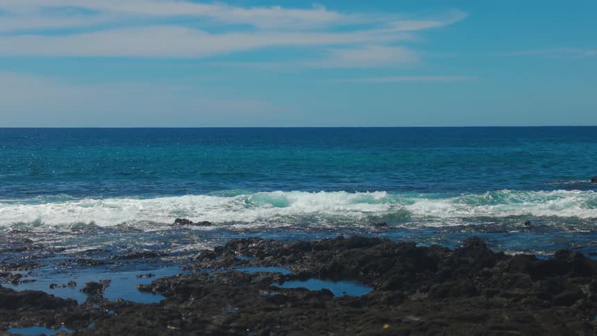 A bright sunny outdoor landscape showing turquoise ocean waves crashing onto black volcanic rock in the foreground under a vast blue sky