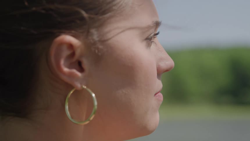 A profile closeup of a young woman wearing a large gold hoop earring with a calm expression looking forward by a body of water with trees in the distant background