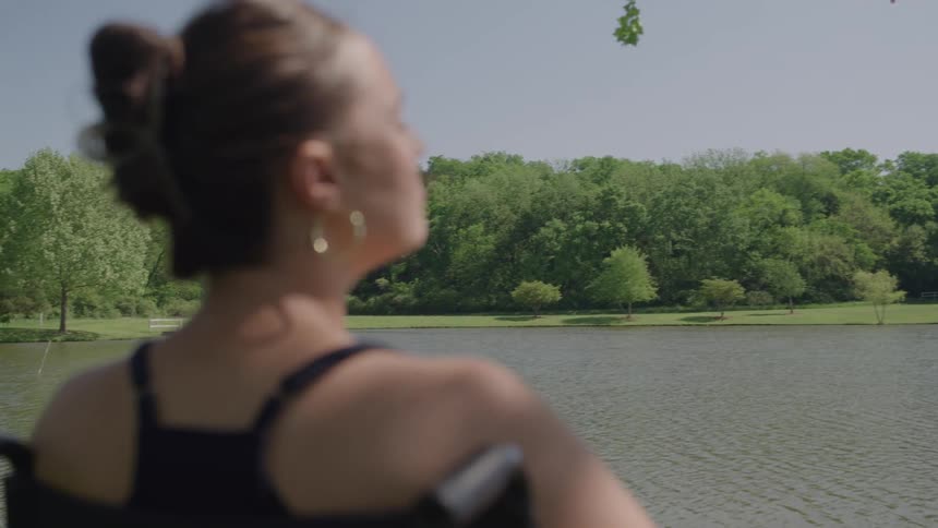 A woman sitting in a wheelchair looks out toward a calm lake bordered by lush green trees on a bright and sunny day