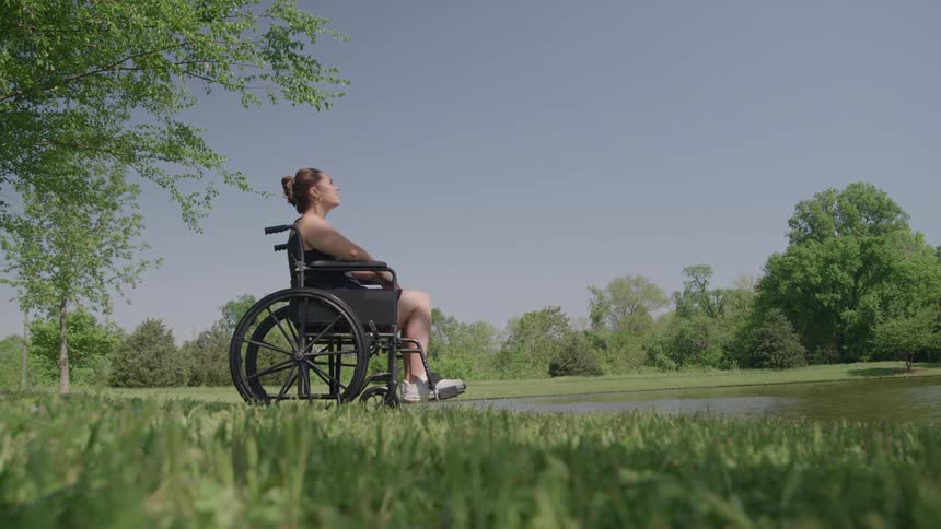 The video features a young woman sitting in a black wheelchair on a patch of grass near the edge of a pond or small lakeshe is dressed casually and slowly raises her head to look up at the bright clear blue sky framed by lush green trees and foliage