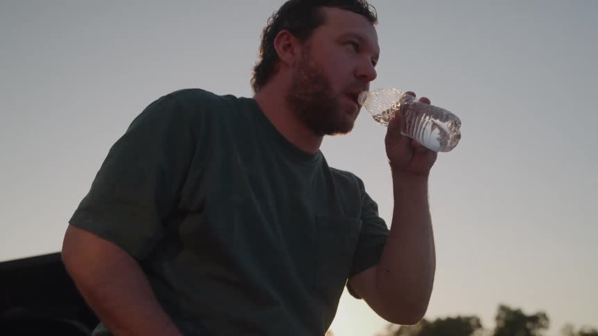 A worker drinks water while resting on a trucks tailgate in golden light at dusk