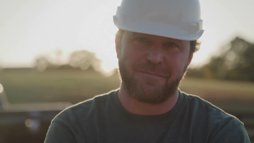 This is a medium closeup shot of a man wearing a white hard hat and a dark green shirt outdoors at sunsethe initially looks down obscured by strong lens flare and sun glare before raising his head to smile slightly at the camera