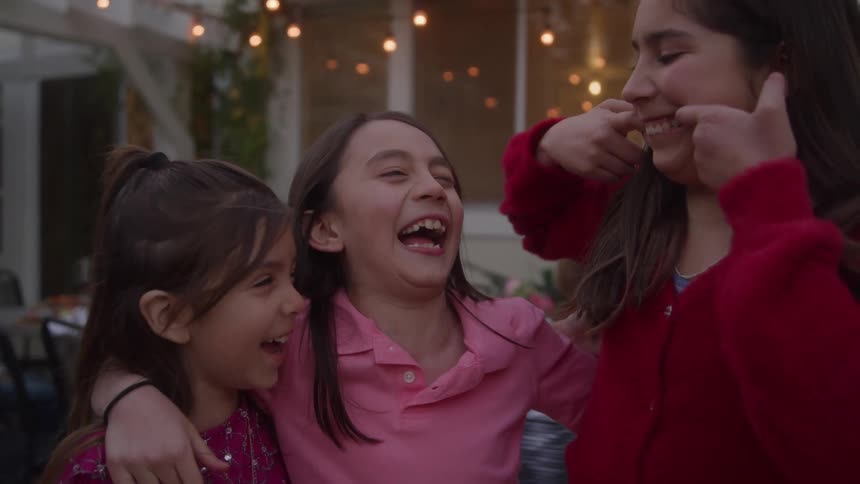 Three young girls are embracing and laughing at silly faces joyously in an outdoor backyard setting with string lights in the background