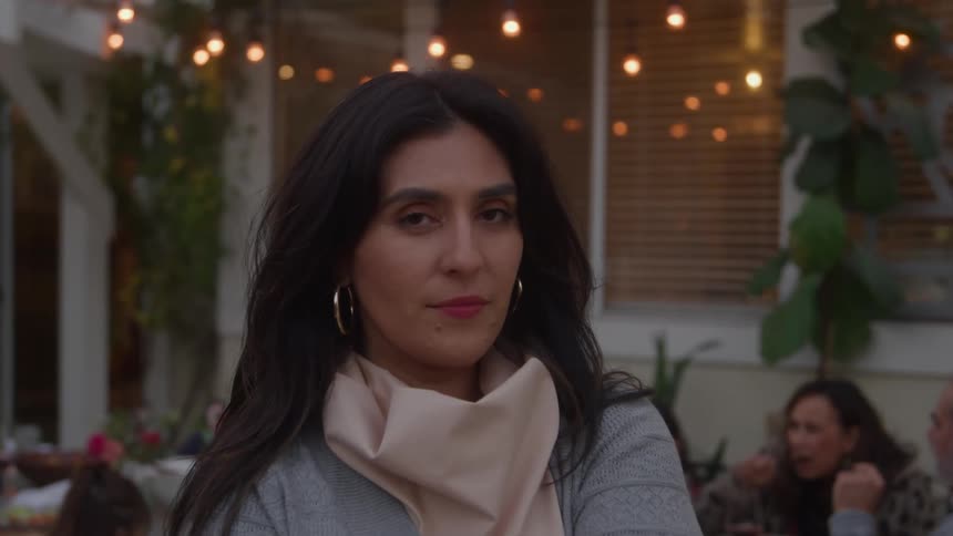 A woman with long dark hair wearing a light grey sweater and a beige scarf looks directly into the camera with a subtle smile in a medium closeup shot set outdoors on a patio with illuminated string lights and blurry figures of people gathering