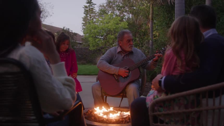 A family gathers around an outdoor fire pit at dusk as an older man plays an acoustic guitar for a small girl while sitting on a patio