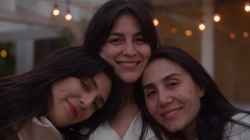 Three adult women with dark hair are captured in a medium closeup smiling warmly and hugging tightly all set against a softly blurred background adorned with glowing string lightsea