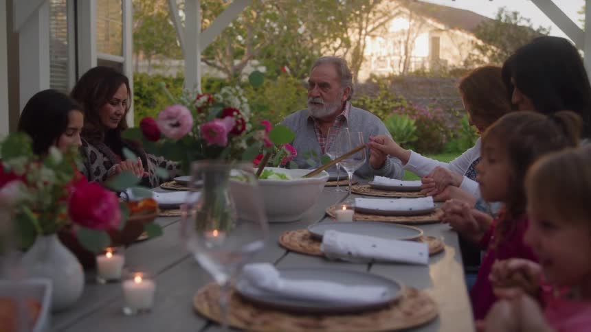 A multigenerational family including an older man and several children sit around an outdoor dining table holding hands and bowing their heads to say grace before eating dinner