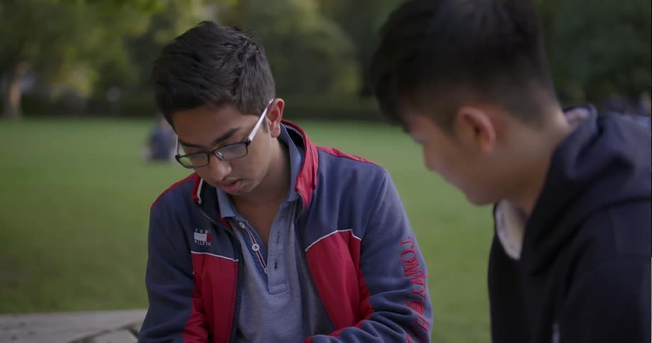 Two students sit on a bench looking over a notebook and talking