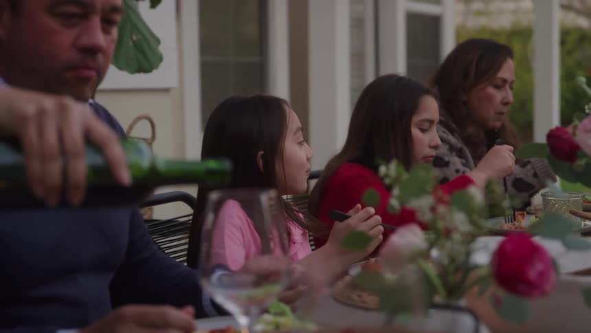 A medium shot shows a family of four dining together outdoors on a patio with a man in the foreground pouring red wine into a glass while two girls and a woman talk and eat in the background