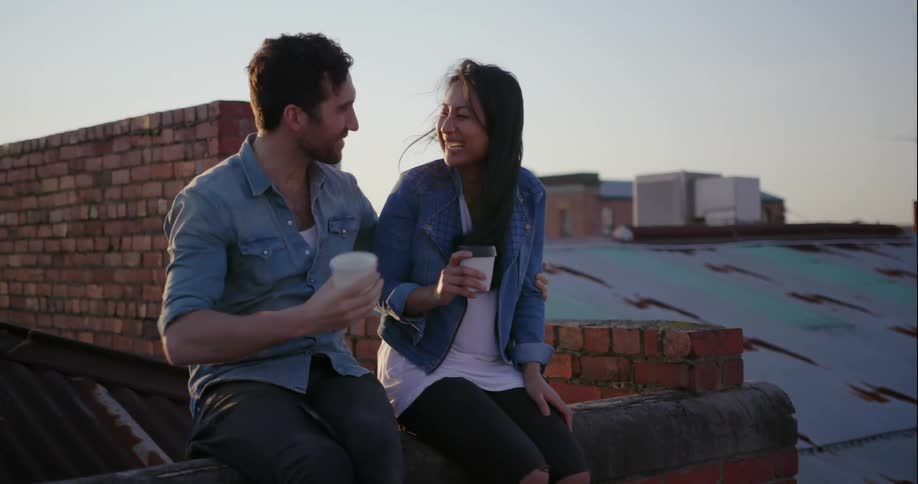 A man and woman laugh and kiss while drinking coffee on a rooftop