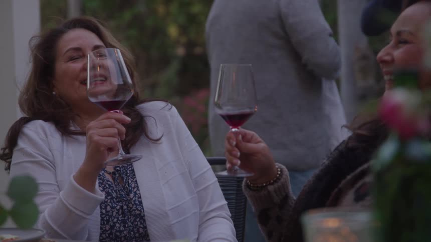 Older women clink wineglasses while sitting at a table outdoors at a friendly get together