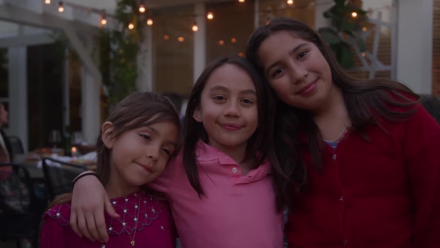 This is a medium closeup of three young girls standing close together smiling and embracing likely siblings or friendsthey are outdoors in what appears to be a backyard or patio setting with strungup lights visible in the blurred background