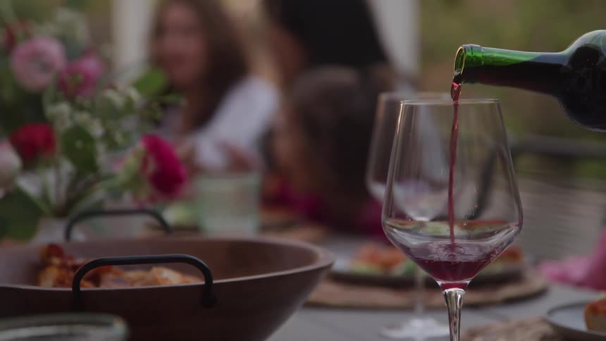 Red wine is poured from a green bottle into a large clear wine glass in the foreground while a blurred group of people are seen enjoying a meal together in the background