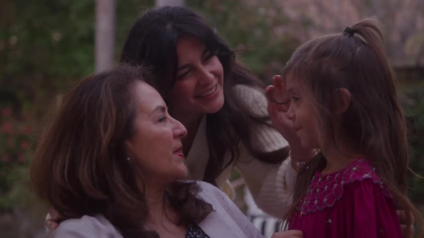Mother and grandmother smile as they interact with a girl while outdoors on an autumn day