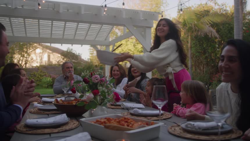 A large family seated at an outdoor table applauds as a happy woman wearing a pink skirt brings a serving bowl to the group during a meal under a white pergola