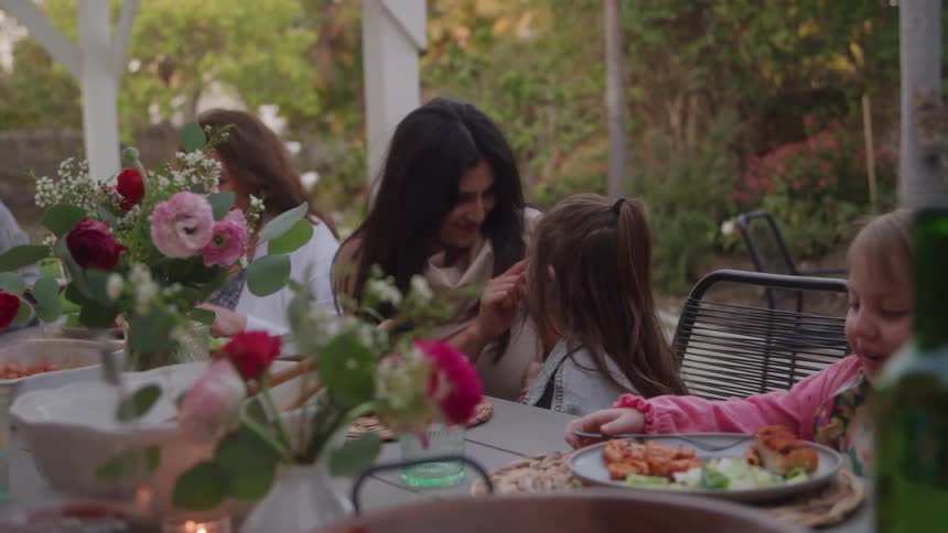 The video shows a family gathered around a table outdoors under a white pergola or canopy enjoying a meal decorated with bouquets of pink and red flowersthe central action features a darkhaired woman interacting affectionately with a young girl sitti