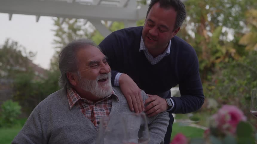 Older man seated outdoors being touched on the shoulder by another man standing beside him