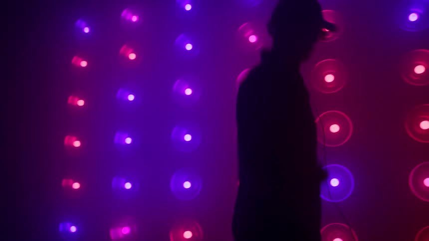 A silhouette of a man wearing a cap and headphones dances rhythmically against a background of flashing purple and blue wall lights