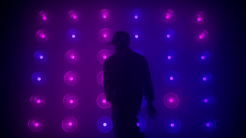 A man is seen in silhouette dancing energetically in front of a grid wall of circular lights that rapidly change color cycling through bright combinations of blue orange red and purple