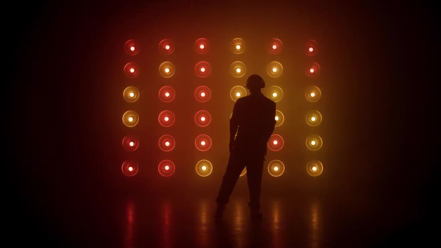 A fullbody silhouette of a dancer wearing a cap and headphones performs dynamic movements against a dramatic completely dark backdrop illuminated by a large grid of lights that flash and change color in rhythmic patterns