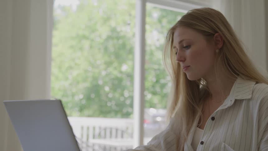 A young woman with long blonde hair dressed in a striped shirt as she smoothly opens a laptop on a reflective glass table in a brightly lit room with a view of greenery outside