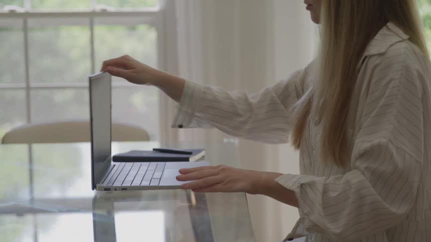 A woman wearing a lightcolored striped shirt seated at a glass table typing on her opened laptop before she deliberately closes the device and stands up in a bright indoor setting near a curtained window