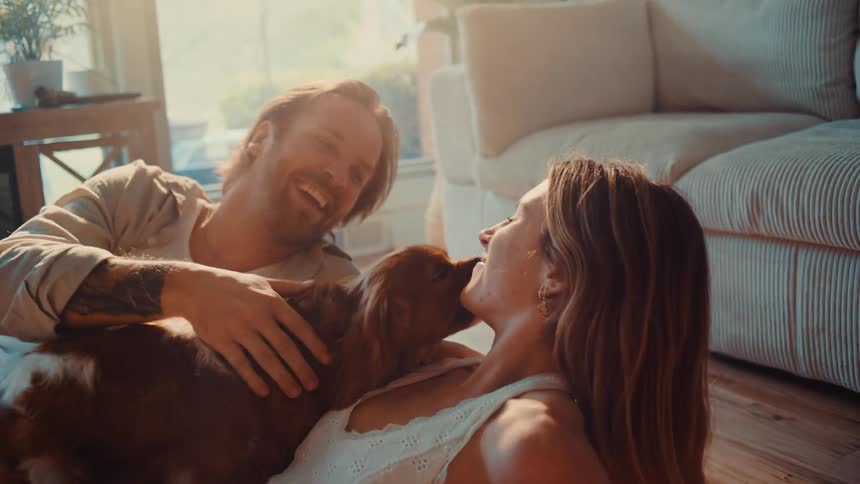 A man and woman are lying on the floor in a sunlit living room with their dog the dog is licking the womans face as the man pets the dog