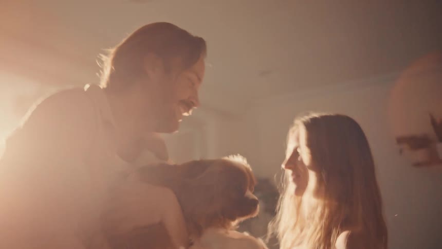 A happy couple dances together holding a small dog in a brightly lit living room