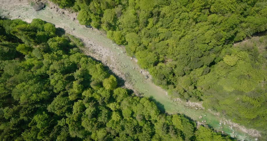 An aerial shot shows a river with pale green water flowing through a forest valley transitioning from barren brown trees to lush green foliage