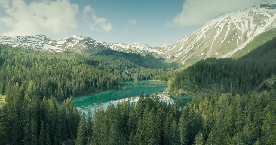 Aerial view of a turquoise lake surrounded by dense forest and snow capped mountains