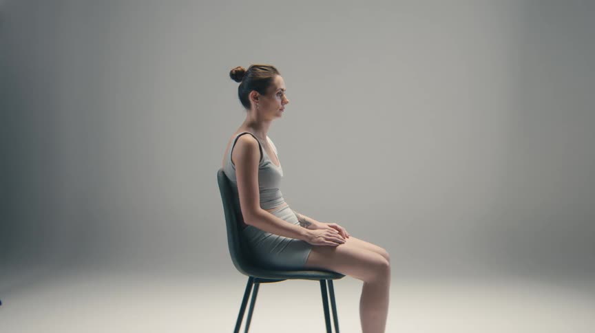A young woman with her hair in a bun sits upright on a modern chair in profile wearing light gray athletic clothing against a smooth gray studio background