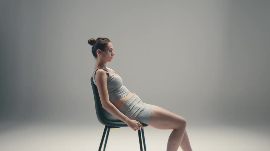 A woman wearing athleisure shorts and a tank top sits in profile on a minimal black chair in a bright neutral studio leaning back against the chairs support in a relaxed contemplative posture
