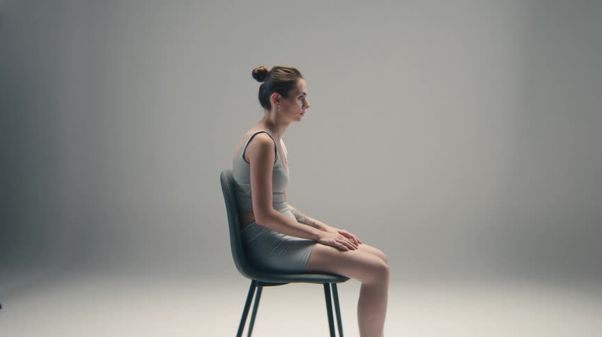 A young woman sits patiently on a chair indoors dressed in innerwear