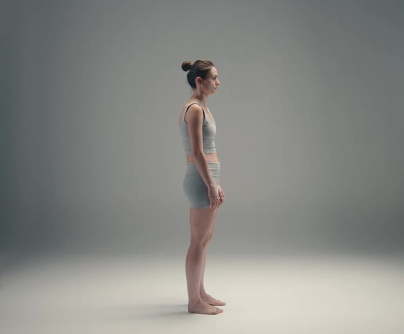 An unsmiling woman in a grey sports bra and shorts stands fully in profile against a simple grey background