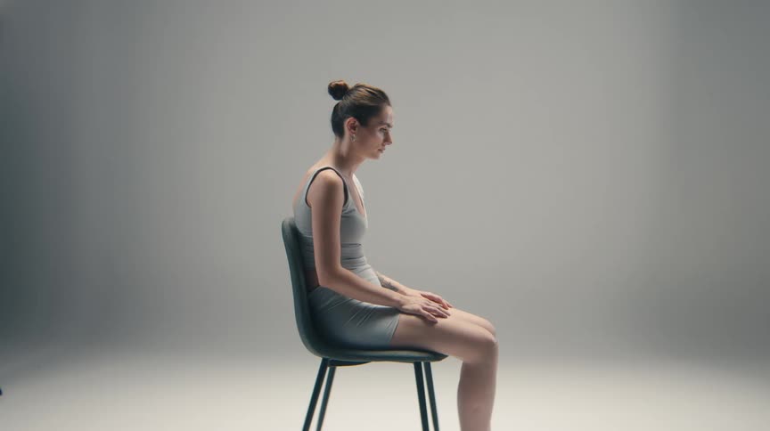 A young woman sits motionless in profile on a chair against a plain grey background