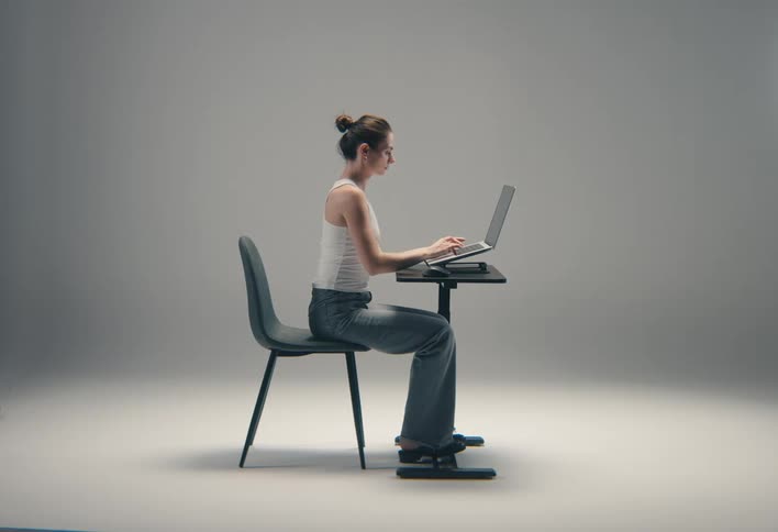 A woman sits in profile at a small desk typing on a laptop with her feet resting on an ergonomic device