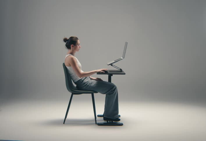 A focused woman in a white tank top and jeans is sitting while working on a laptop at a small table against a grey background