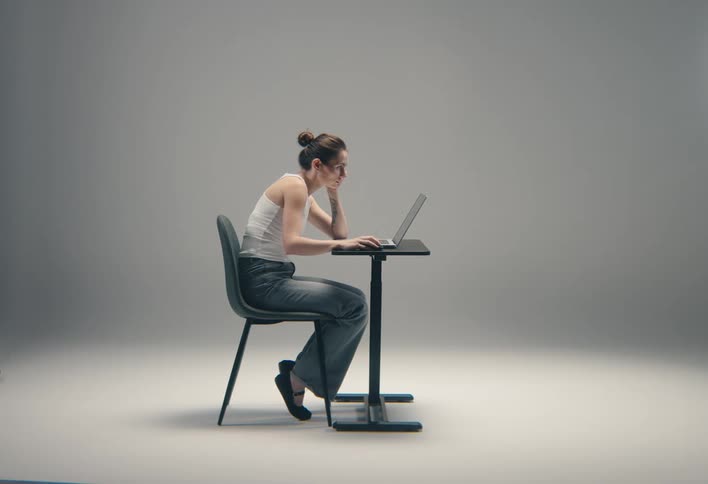 A focused woman in a white tank top and jeans is sitting while working on a laptop at a small table against a grey background