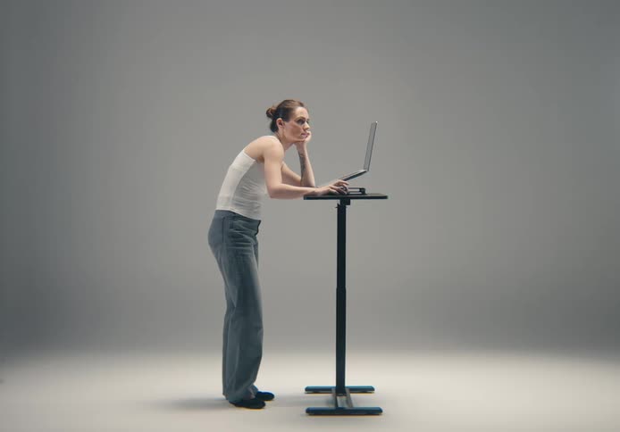 A woman with hunched posture leans on her hand while looking at a laptop on a standing desk