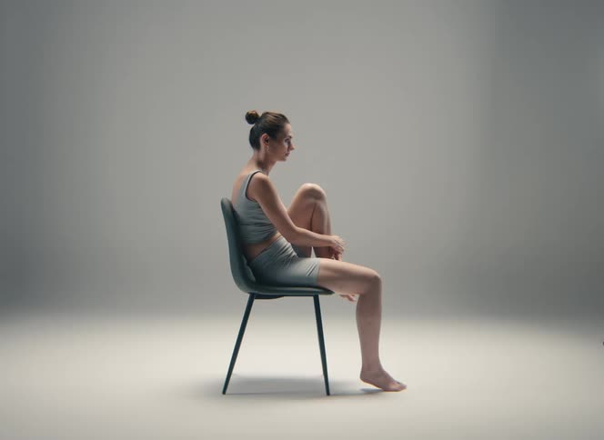 A serious female figure in athletic wear sits on a chair in a bright studio with a grey background