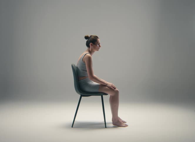 A woman sits on a chair in a studio with her back hunched demonstrating poor posture