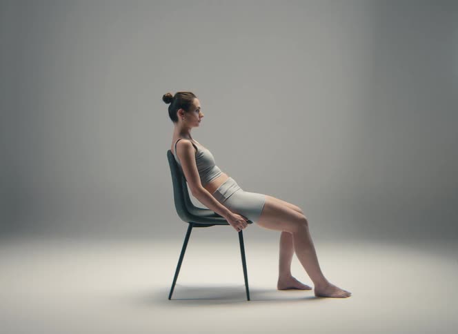 A woman in athletic wear sits with slumped posture on a chair in a plain studio