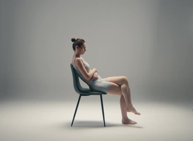 An unsmiling woman in a grey sports bra and shorts is sitting with her arms crossed against a simple grey background